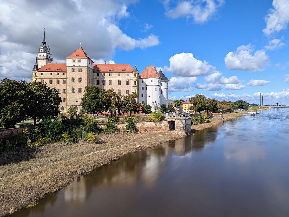 Schloss Hartenfels in Torgau, Sachsen, mit markanten Türmen und roten Dächern am Ufer der Elbe, umgeben von Bäumen und locker bewölktem, blauen Himmel